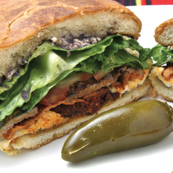 A Mexican torta sandwich, cut in half, revealing layers of meat, lettuce, and cheese, with a jalapeño.